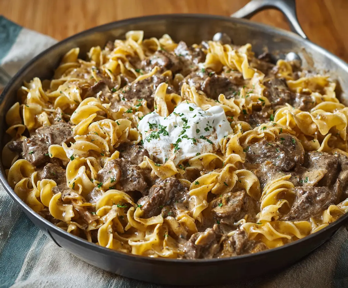 Creamy beef stroganoff served over noodles, garnished with fresh herbs on a white plate.